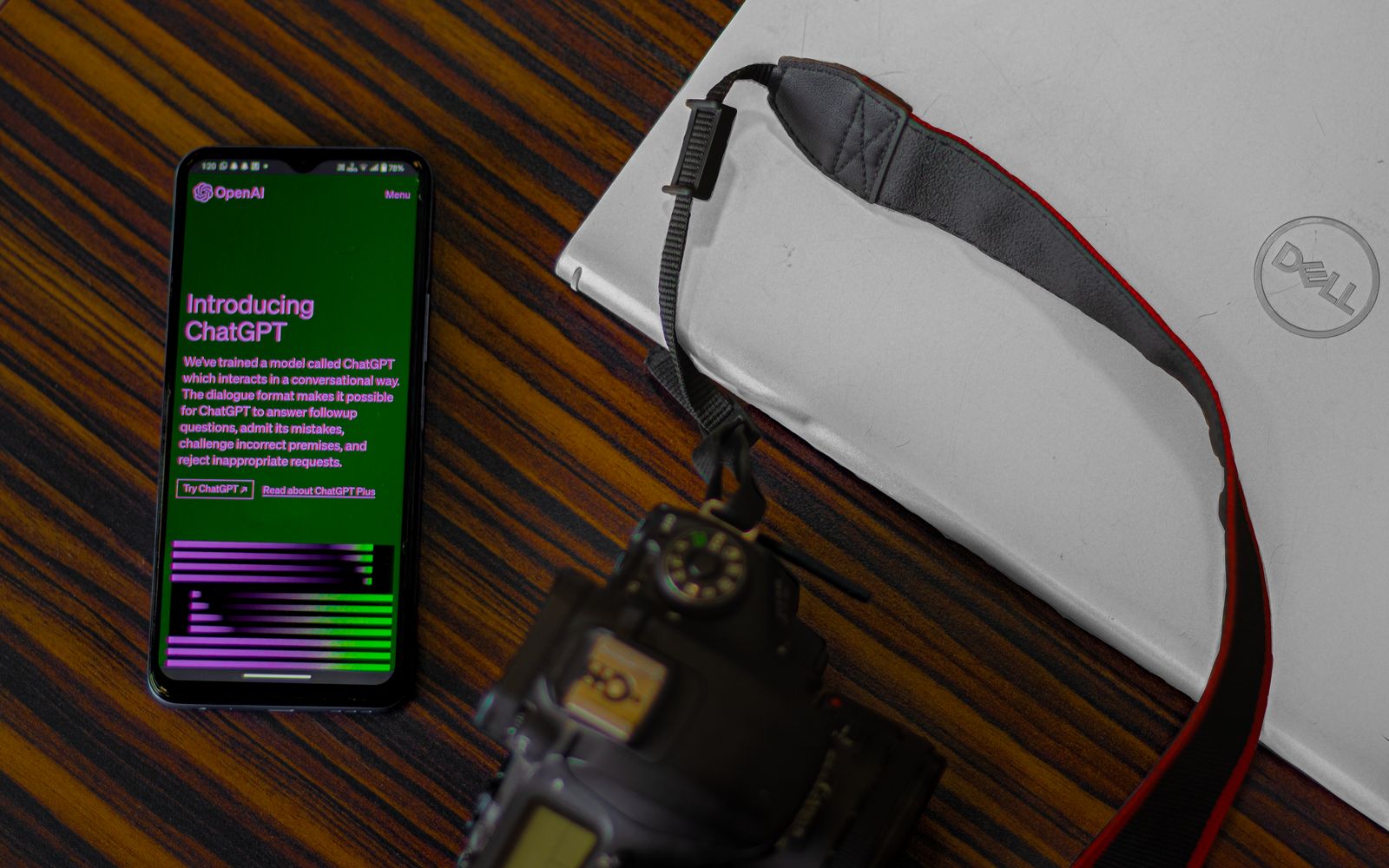 Smartphone with ChatGPT screen next to camera and laptop on wooden desk.