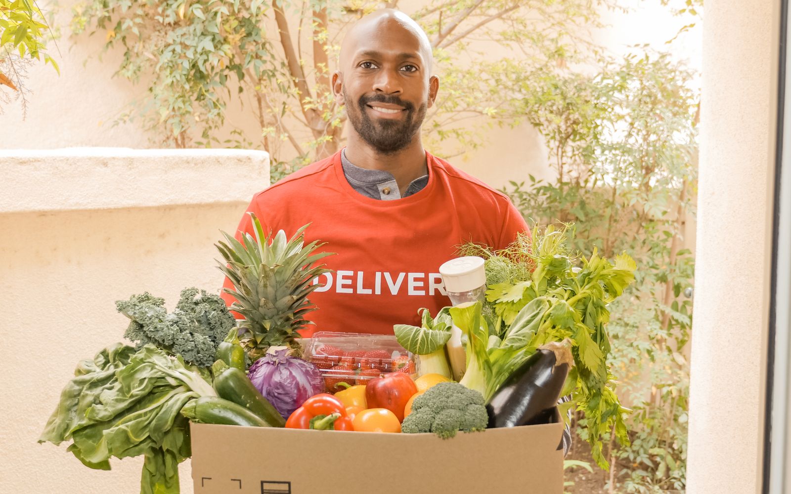 Deliveryman in red shirt holding a box of fresh vegetables, smiling at the doorway.